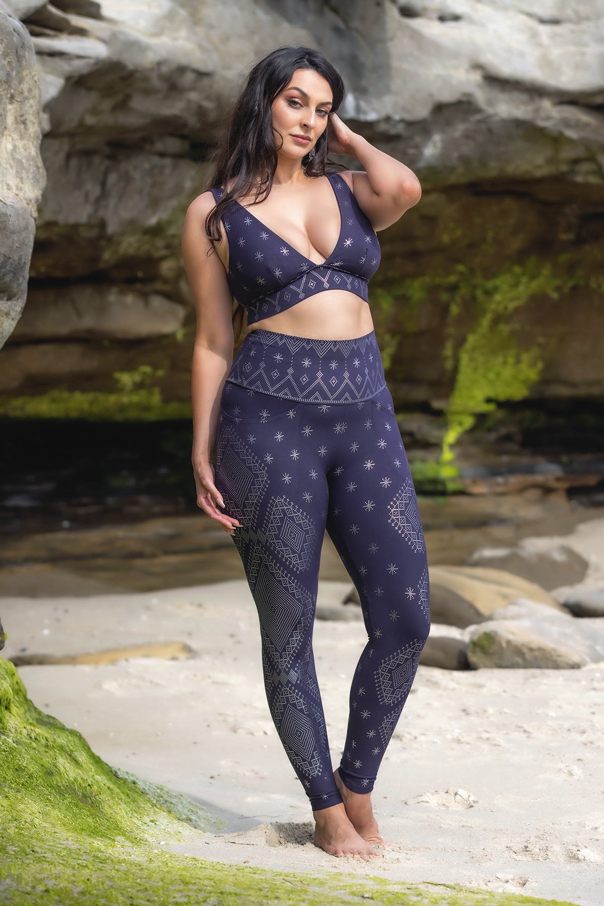 A woman with long dark hair poses on a sandy beach near mossy rocks, wearing the Melodia Designs Dendera Vira Bra Midnight Silver paired with matching leggings featuring geometric and star patterns.