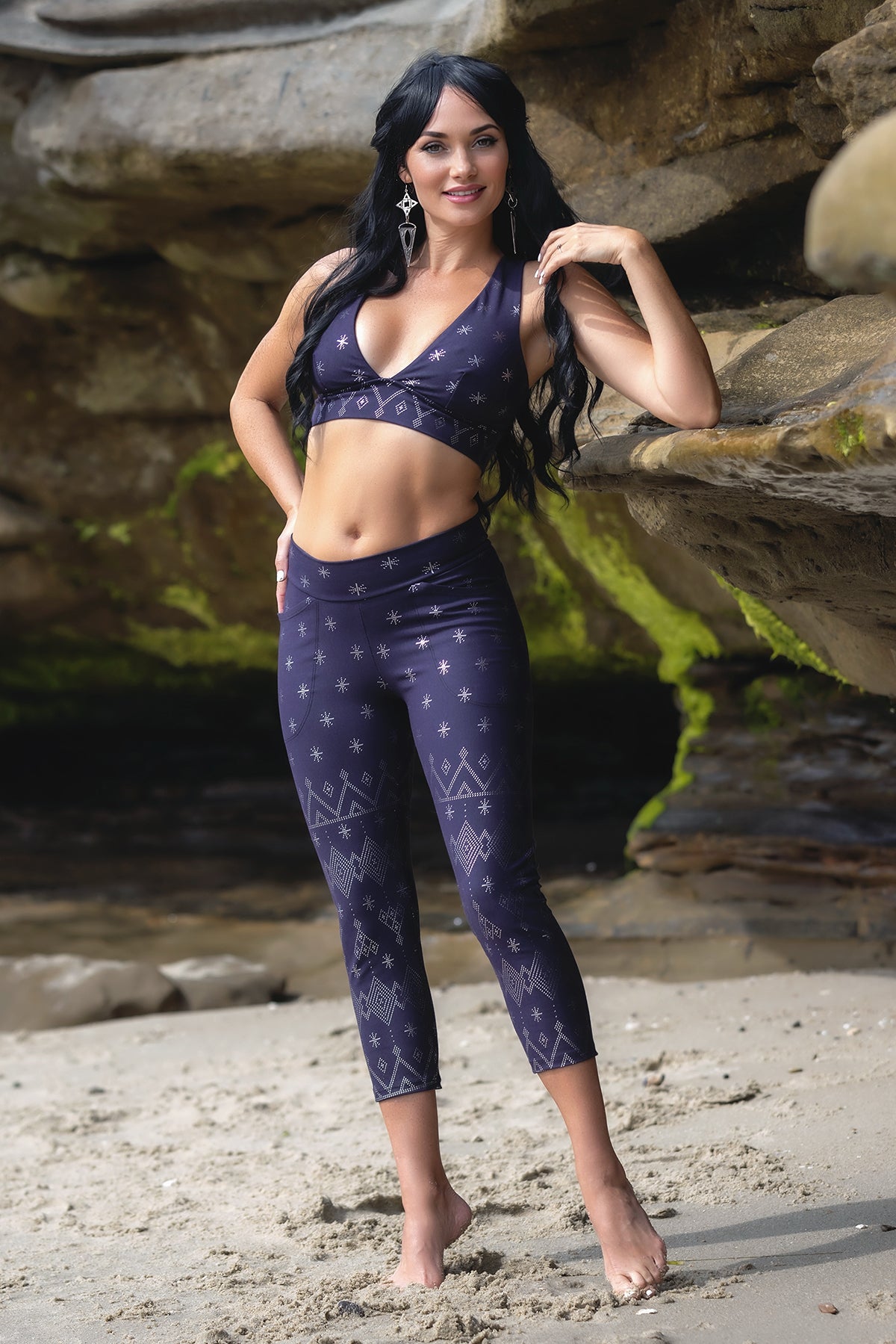 A woman with long black hair wears the Melodia Designs Dendera Zoe Capri Midnight Silver set as she poses barefoot on a sandy beach near mossy rocks.