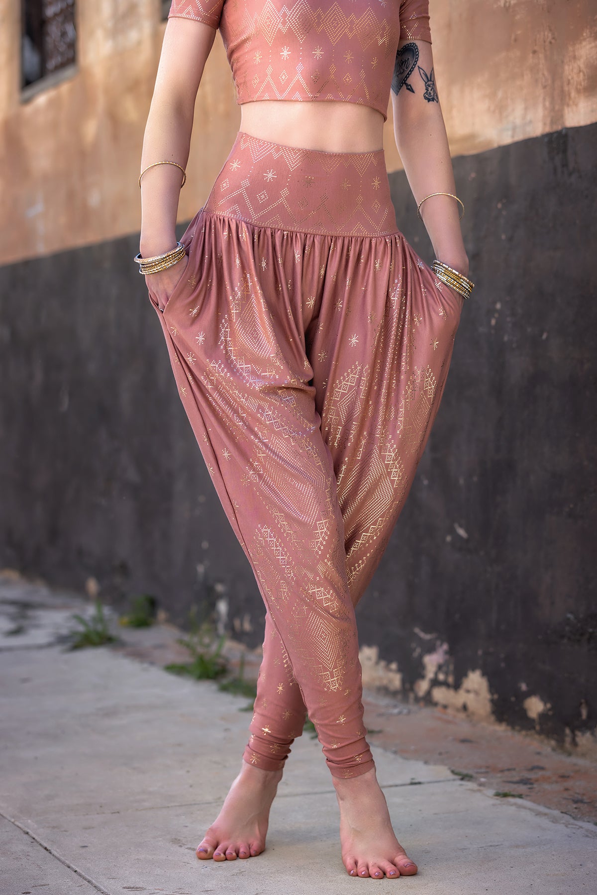 A person wearing Melodia Designs' Dendera Zaina Harem 2.0 Desert Rose Gold set stands barefoot on a sidewalk, hands in pockets, in front of a textured wall.