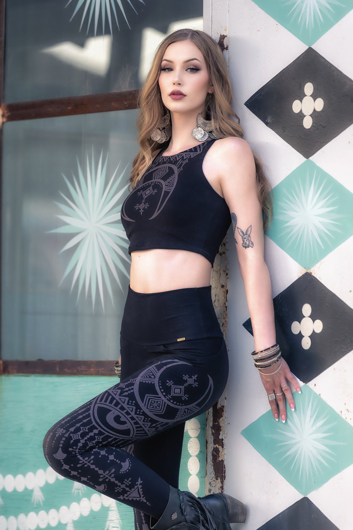 A woman with long hair wears the Melodia Designs Desert Oracle Laura Crop Top and patterned leggings in a striking Desert Oracle print as she leans against a wall with geometric designs, looking confidently at the camera. She has a tattoo on her forearm and wears multiple bracelets.