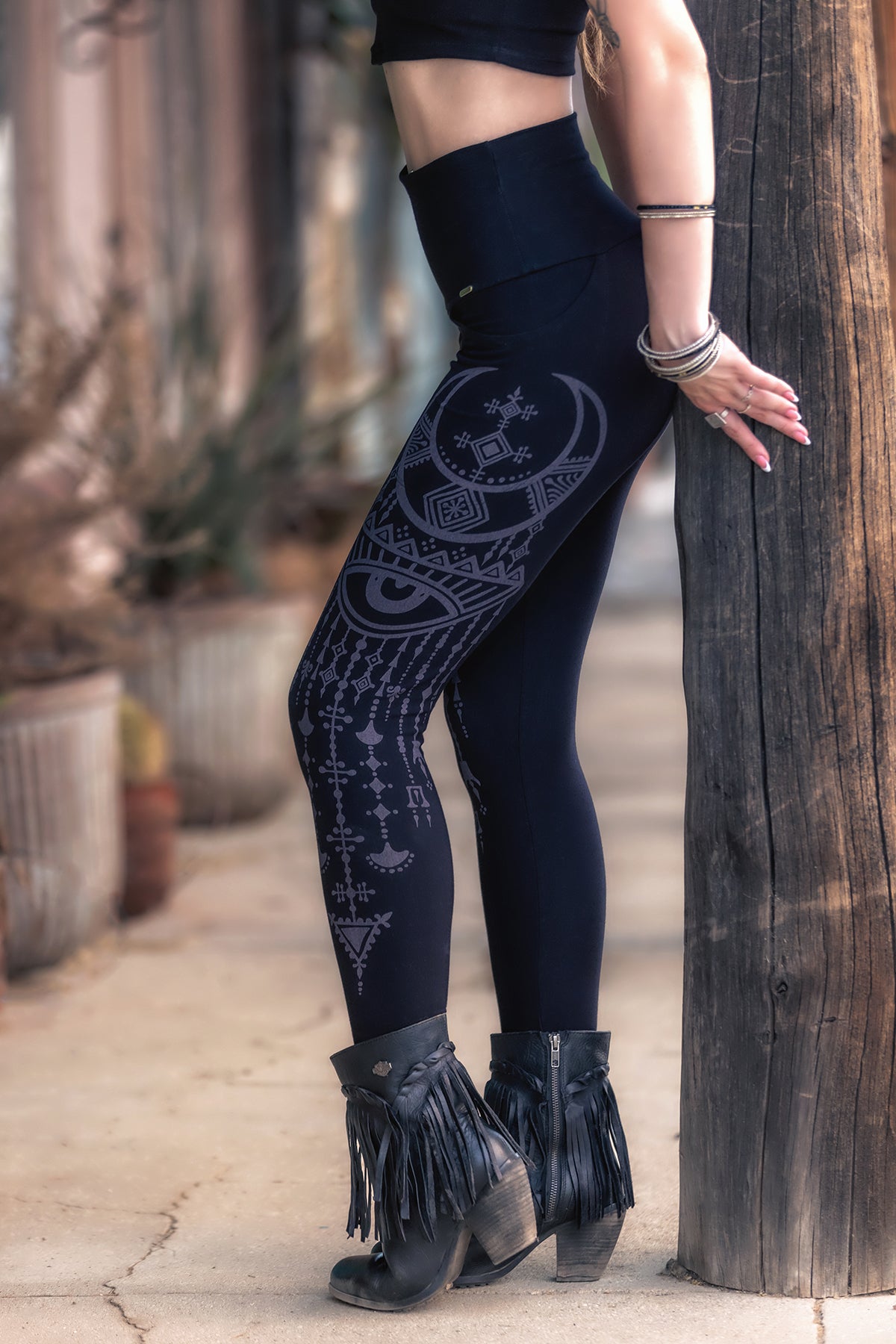 A woman stands outdoors, leaning on a wooden post. She wears Melodia Designs' Desert Oracle Elsa Legging—an eco-luxe bamboo high-rise style featuring the distinctive Desert Oracle print—paired with a black crop top and black fringed heeled ankle boots. Her face is not visible.