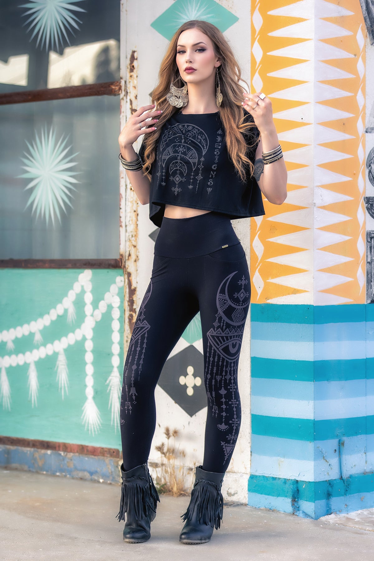 A woman with long, wavy hair poses confidently outside against a vibrant, geometric wall, wearing the Melodia Designs Desert Oracle Dede Crop Top made from eco-friendly modal jersey, paired with matching patterned leggings, fringe boots, and large earrings.