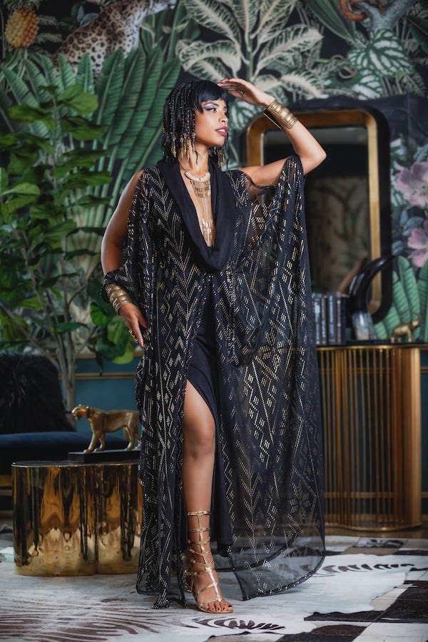 A woman poses indoors in the Melodia Designs Domina Gown, a glamorous black and gold patterned dress, surrounded by lush tropical decor and gold accents, one hand shielding her eyes as she gazes into the distance.