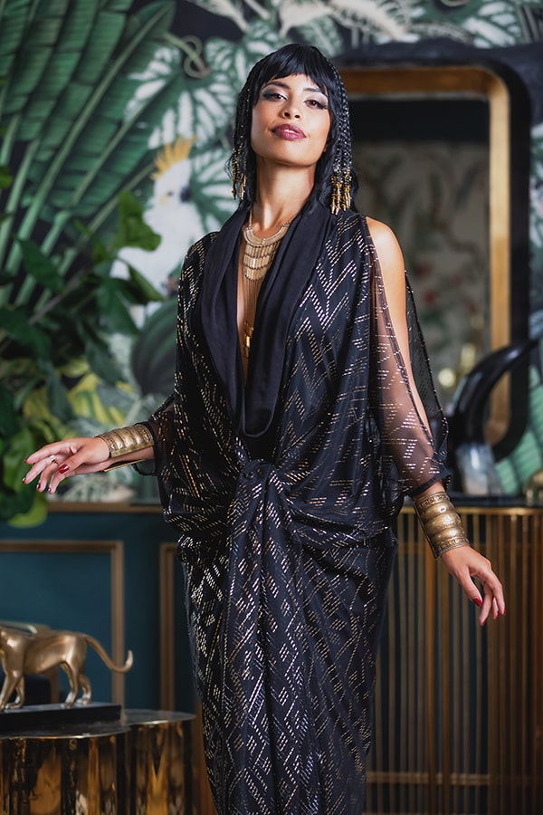 A woman exudes vintage glamour in the Domina Gown by Melodia Designs—its black and gold pattern and her gold jewelry shining amid lush green plants and elegant décor, reflecting her regal, confident presence.