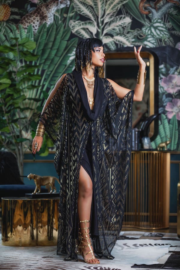 Wearing the Domina Gown by Melodia Designs—a black and gold patterned dress with a versatile silhouette—a woman adorned in gold jewelry poses indoors among tropical plants and animal-themed décor.