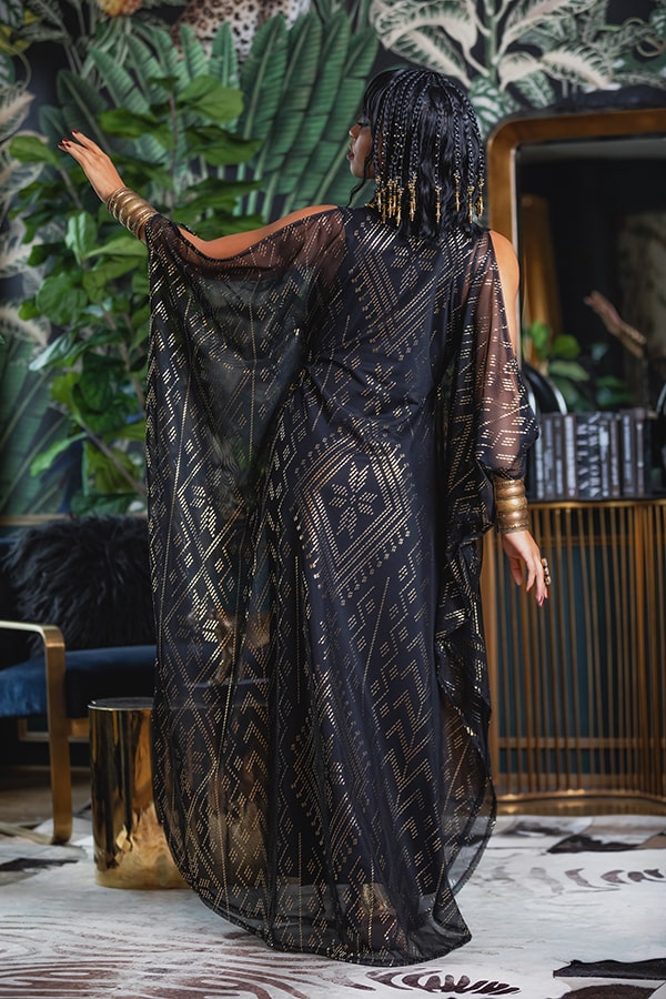 A woman with beaded hair stands with her back to the camera, wearing the Domina Gown by Melodia Designs—a chic black and gold sheer cover-up—in a stylish room filled with leafy plants, a mirror, and modern decor.