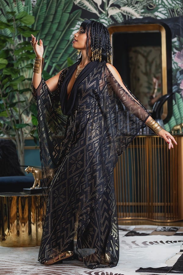 A woman in the Domina Gown by Melodia Designs—a black and gold geometric-patterned sheer dress with a versatile silhouette—poses indoors among lush plants and gold accents, her beaded hair and gold bracelets completing an elegant, regal look.