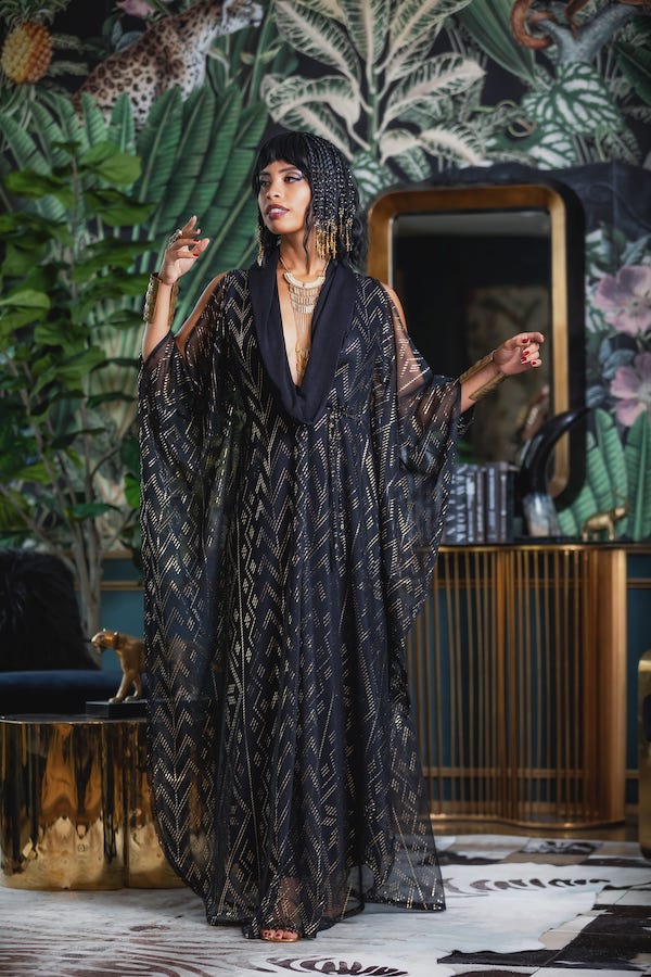 Wearing the Domina Gown by Melodia Designs—a sheer black and gold geometric caftan—a woman channels vintage glamour indoors, set against tropical wallpaper, gold decor, lush green plants, layered jewelry, and a beaded headdress.