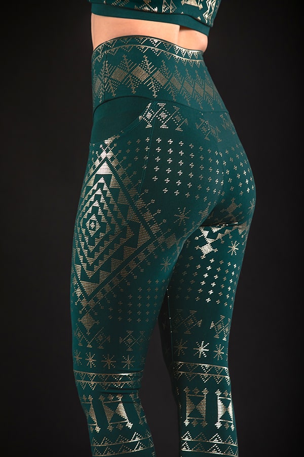 A person wears Melodia Designs Alkimiya Elsa Pocket Legging Malachite—full-length, dark green with a high-rise foldable waistband and shiny gold geometric and snowflake patterns—standing against a black background.