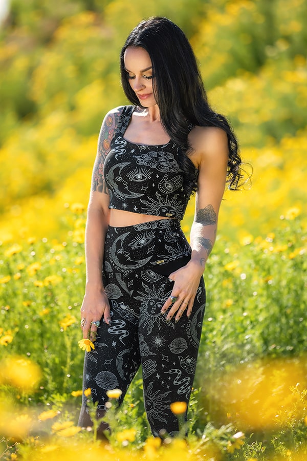 A woman with long dark hair and tattoos wears the Neisha Bra Celestial by Melodia Designs—a black cropped tank with white celestial patterns—while standing in a sunny field of yellow flowers.