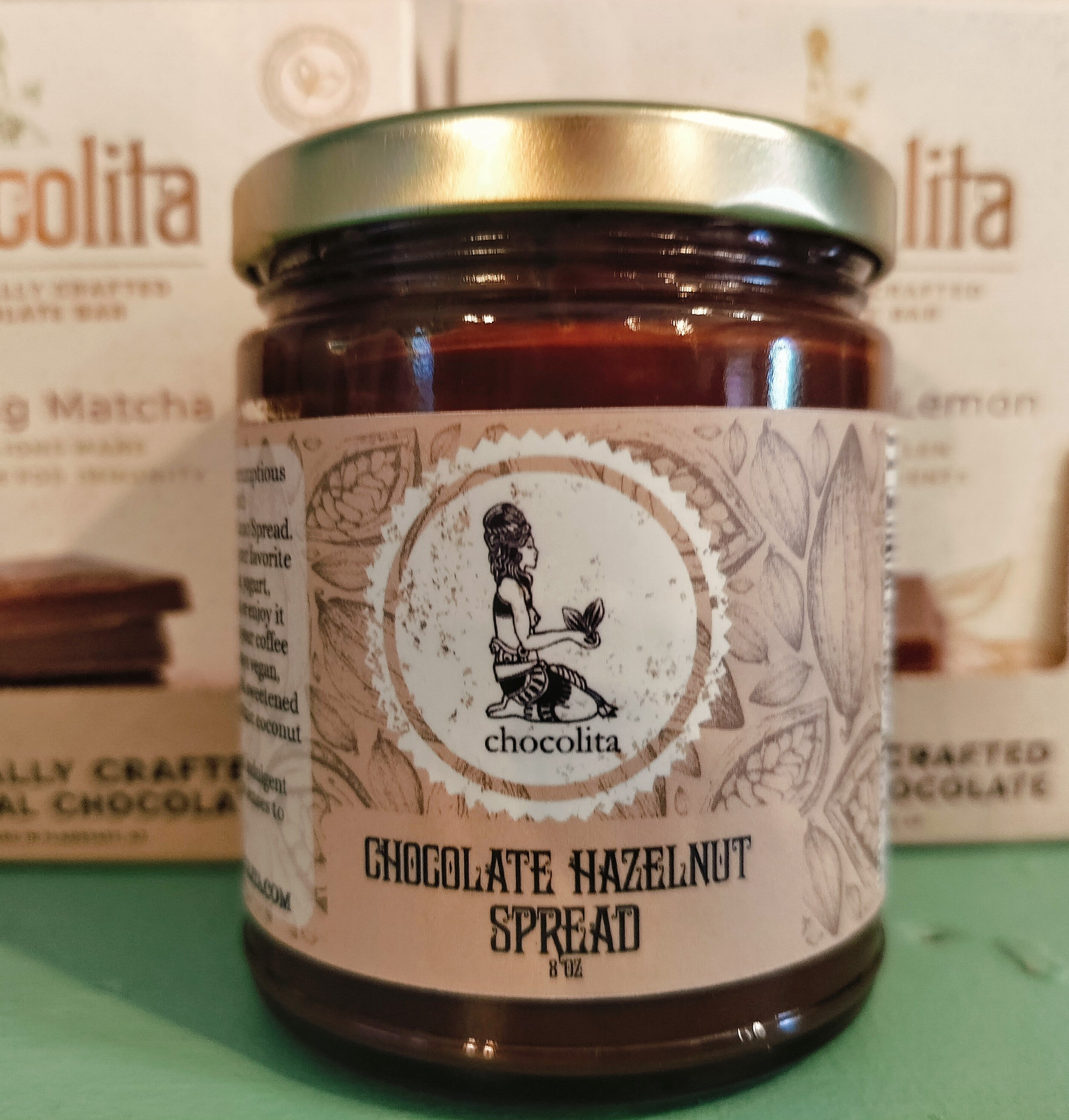 A jar of chocolita.com Hazelnut Chocolate Spread with a label of a meditative figure among cocoa pods and leaves sits in front of chocolita.com chocolate boxes.