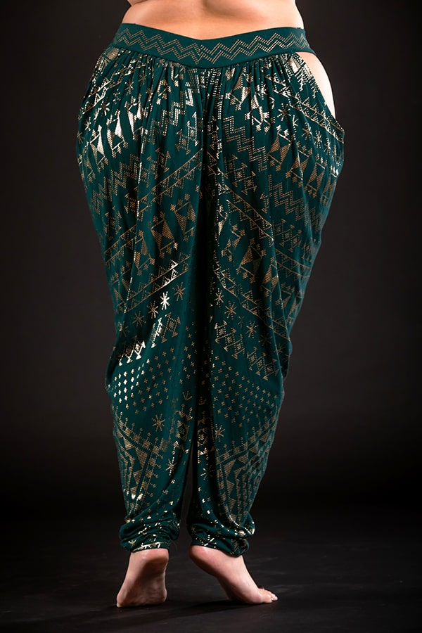 Barefoot, a person wears Melodia Designs Alkimiya Isha Harem Pant Malachite—loose, eco-luxe bamboo pants in dark green with metallic gold geometric and star patterns on a black background.