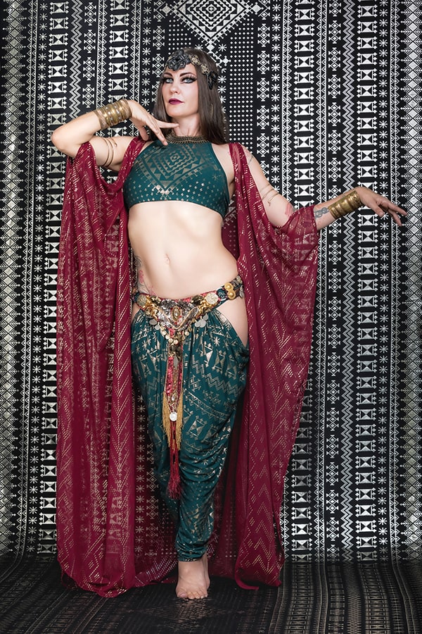 A woman in a green and gold costume, wearing Melodia Designs Alkimiya Isha Harem Pant Malachite and a maroon sheer shawl, poses gracefully before a black-and-white geometric backdrop, adorned with jewelry and a headpiece.
