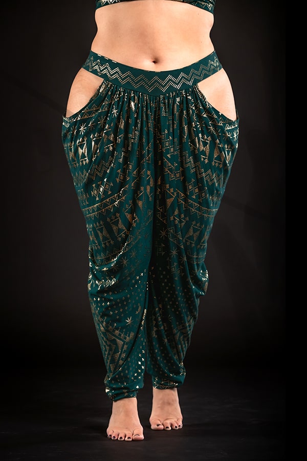 A person models Melodia Designs’ Alkimiya Isha Harem Pant Malachite—eco-luxe bamboo pants with gold geometric patterns, a wide waistband, and open hip cutouts—barefoot against a black background.