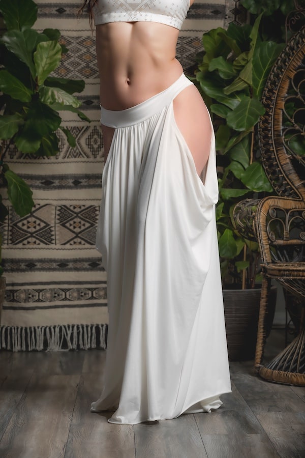 Someone wears the Melodia Designs Aurora Peekaboo Palazzo Ivory, featuring high side slits, indoors on a wooden floor with green plants and woven decor in the background.