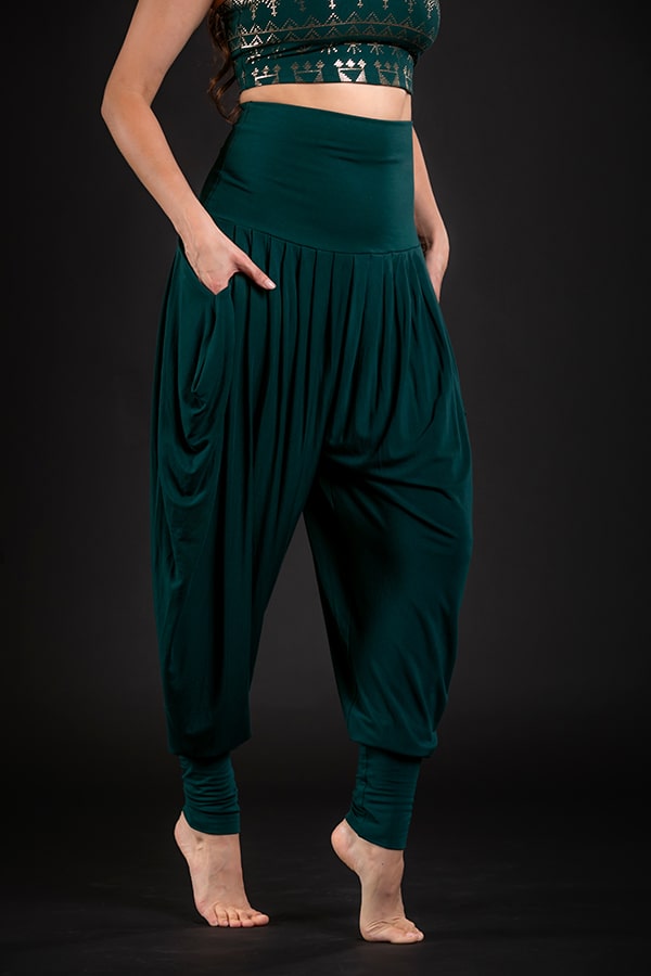 A person poses barefoot in Melodia Designs Jazmine Harem Pant Malachite, featuring a fold-over waistband and deep side pockets, paired with a matching crop top against a black background. The face is hidden—an eco-luxe essential look.