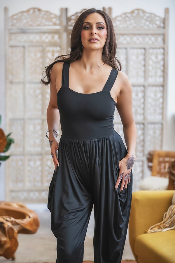 A woman with long dark hair wears the Melodia Designs Jinni Jumper, a sleeveless black harem pant style, standing indoors with one hand in her pocket. Decorative wooden screens and a yellow chair appear in the background.