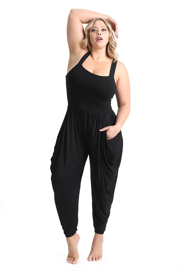 A woman poses barefoot in the Melodia Designs Jinni Jumper— a stylish black, sleeveless jumpsuit with loose harem-style pants—standing against a plain white background.