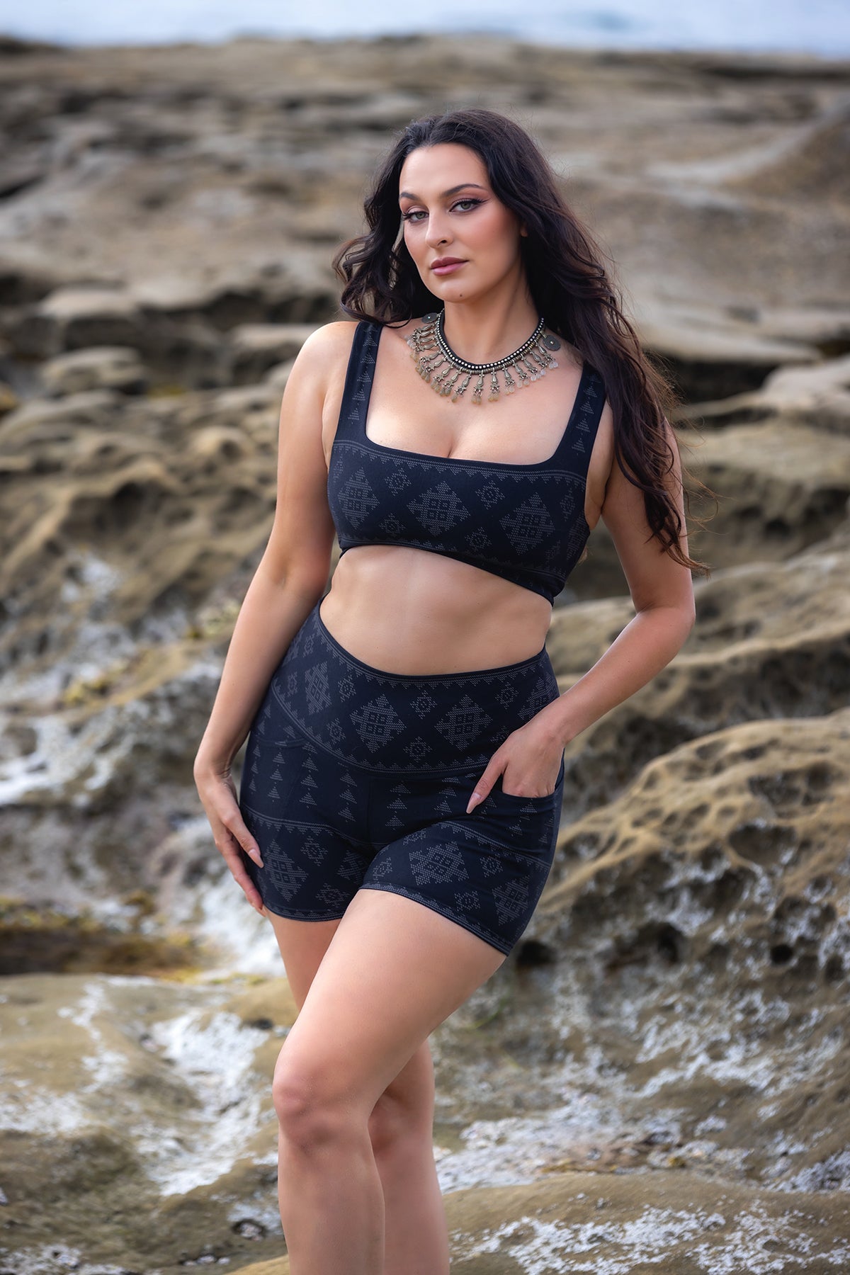 A woman with long dark hair poses on rocky terrain, wearing Melodia Designs' Karnak Alice Short Shadow with a high waistband and geometric patterns, paired with a matching dark sports bra and a statement necklace.