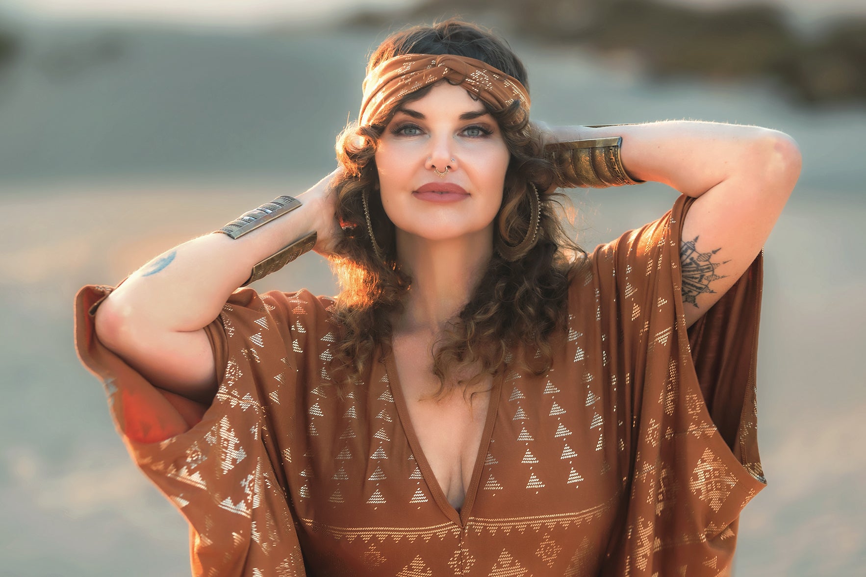 A confident woman with curly hair and a patterned brown dress wears Melodia Designs’ Karnak Beauty Band Copper on a sandy beach, gold foil bracelets adorning her wrists, hands behind her head as she gazes at the camera.