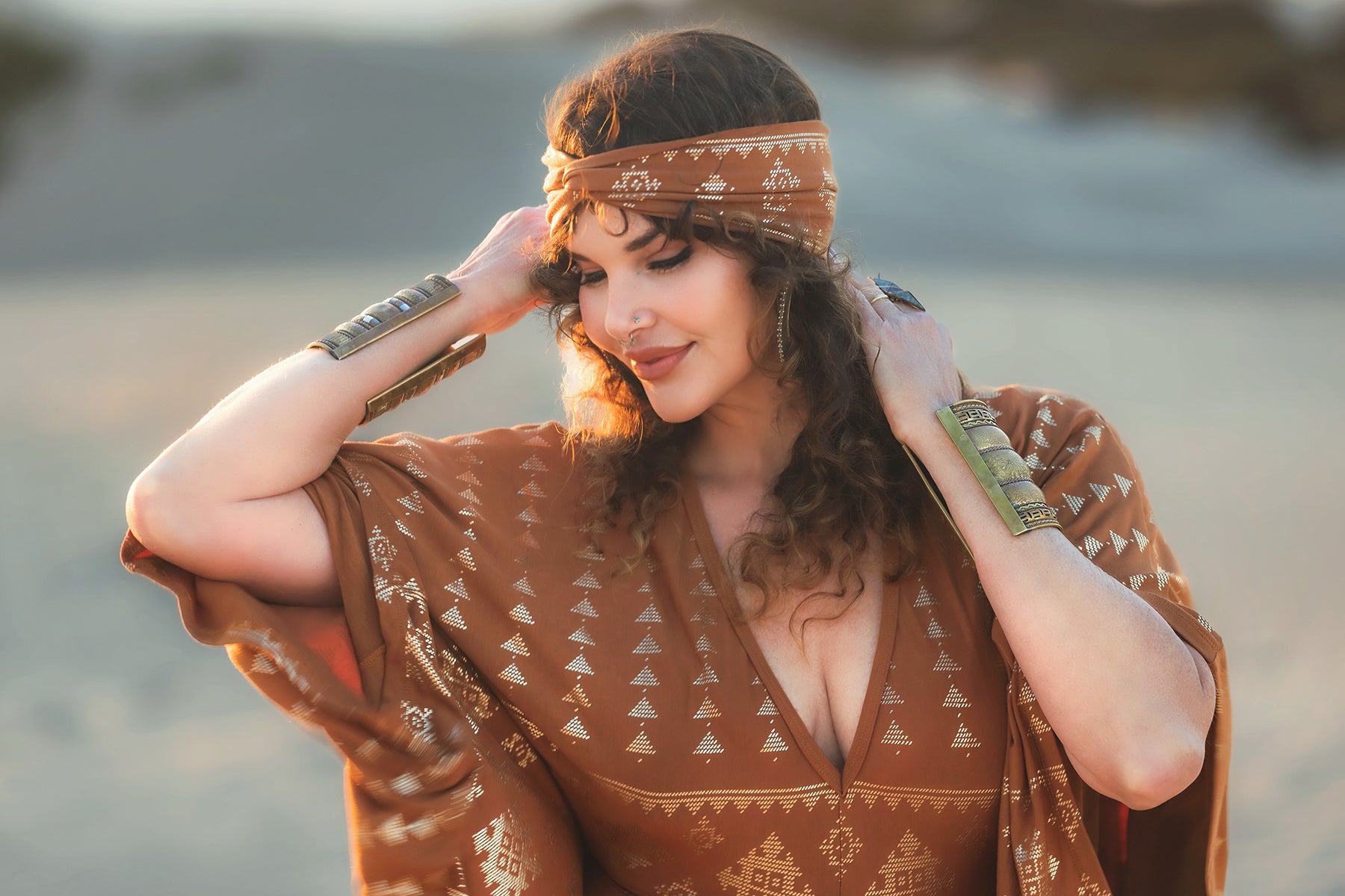 A woman in a patterned brown dress and the Karnak Beauty Band Copper by Melodia Designs stands outdoors, smiling softly with her eyes closed. Her curly hair is elegantly styled, and she wears metallic bracelets with a blurred natural background behind her.