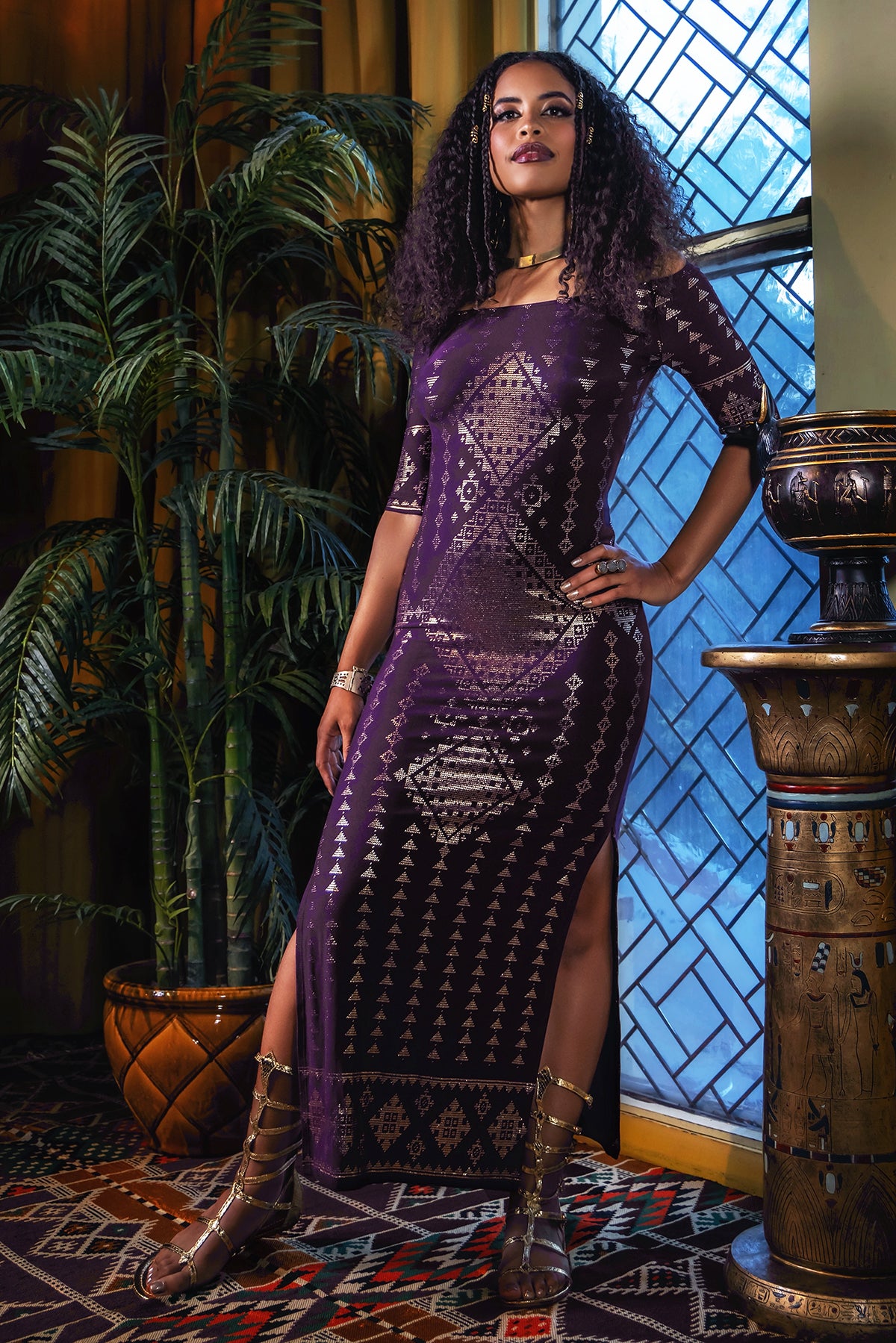 A woman with curly hair wears the Karnak Beledi Dress Aubergine by Melodia Designs—an elegant, long purple patterned dress with side slits—paired with gladiator sandals. She stands confidently indoors beside a tall plant and geometric windowpanes.