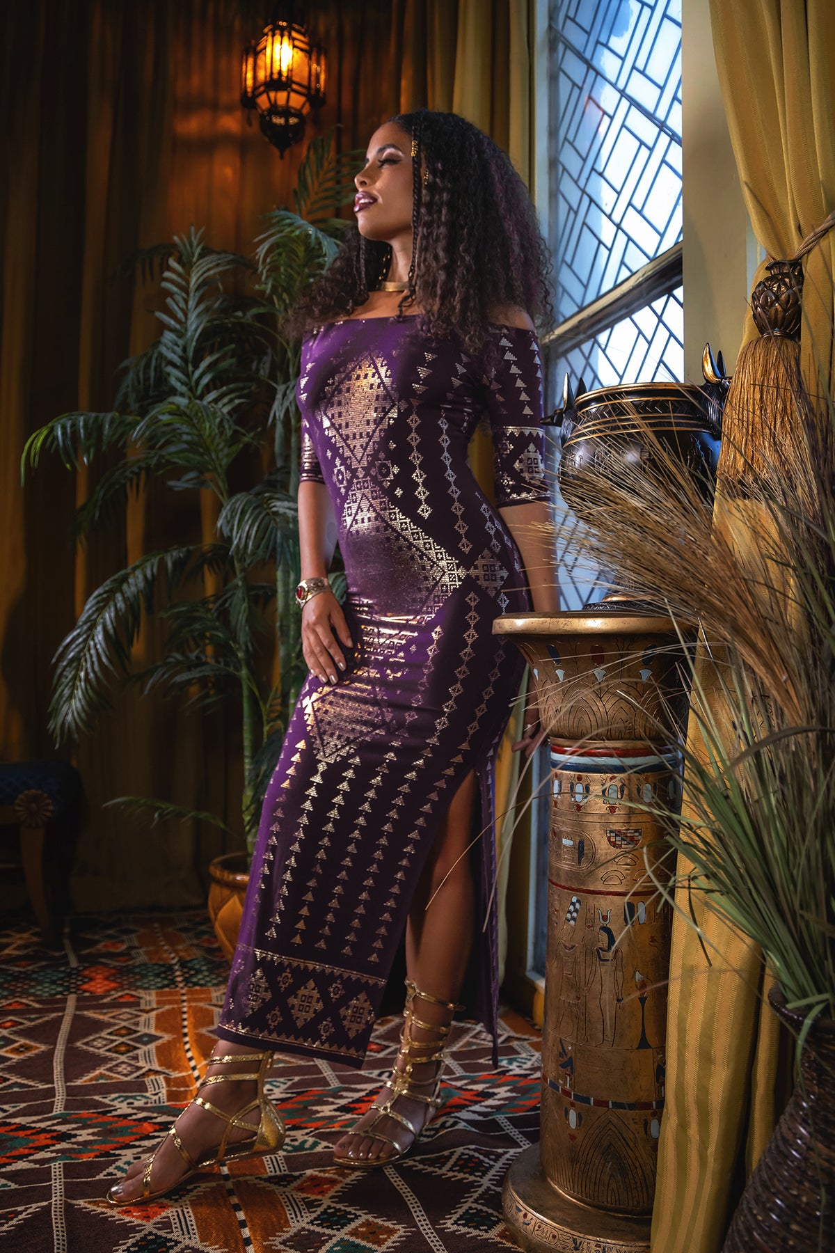 A woman in the Melodia Designs Karnak Beledi Dress Aubergine-Gold stands by a window in a warmly lit room with gold curtains, Egyptian decor, and plants. She accessorizes with gold sandals and jewelry, exuding a relaxed, confident vibe.