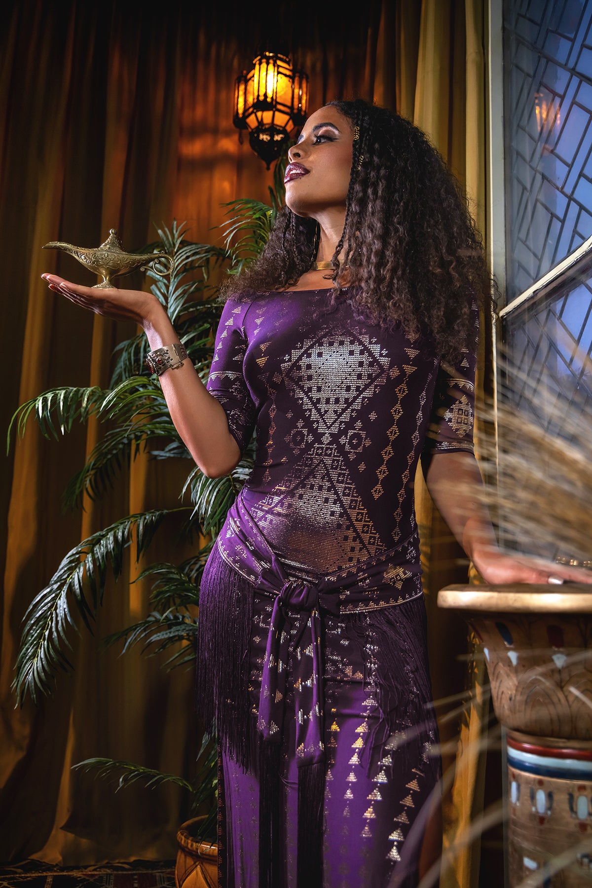 A woman in the Karnak Beledi Dress Aubergine-Gold by Melodia Designs holds a golden lamp near a potted plant and geometric window, under warm lighting.