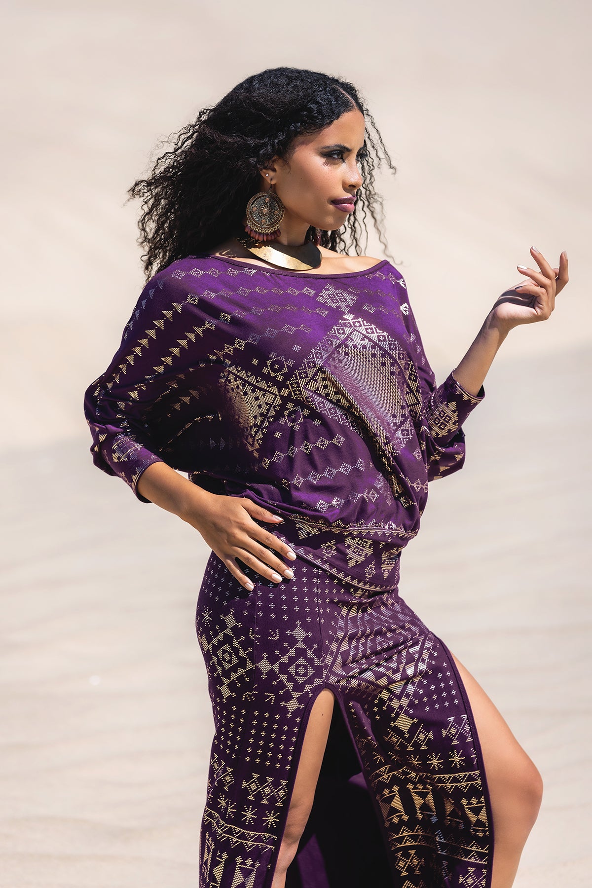A woman in Melodia Designs' eco-chic Karnak Bella Batwing Blouse in aubergine stands on sand, looking to the side. She wears large earrings, has curly hair, and raises one hand with effortless grace.