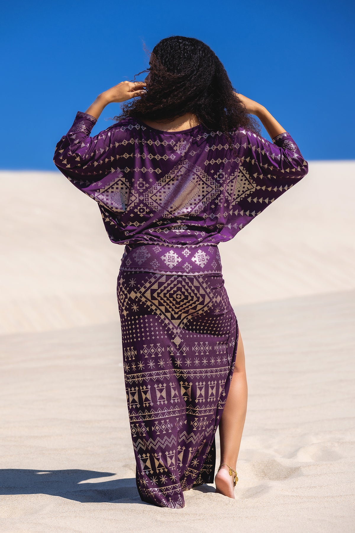 A person with long curly hair stands barefoot on sand, facing away, wearing the Melodia Designs Karnak Bella Batwing Blouse in Aubergine, featuring a flowing silhouette and gold print, beneath a vivid blue sky.