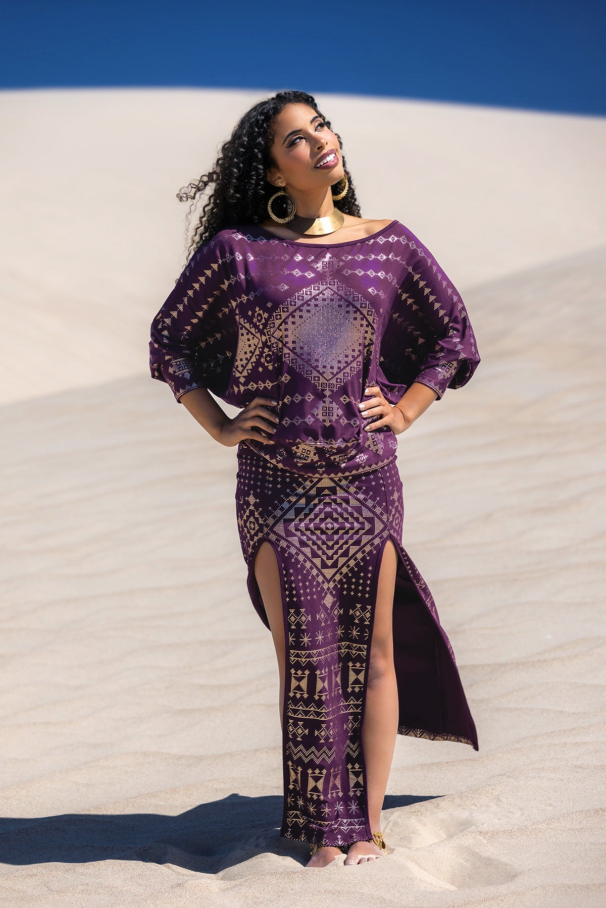 A woman with curly hair stands on sandy dunes, smiling under a clear blue sky. She wears Melodia Designs’ Karnak Bella Batwing Blouse in Aubergine with geometric print and side slits, paired with gold hoops and a statement necklace.
