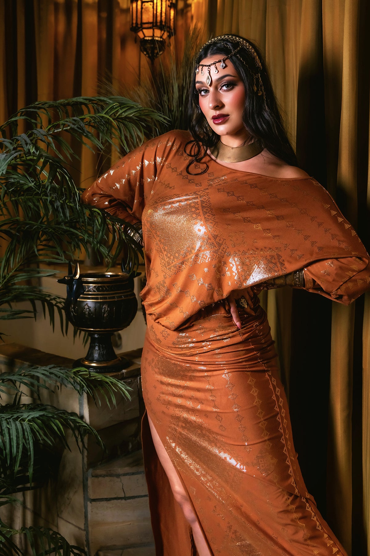 A woman models the Melodia Designs Karnak Bella Batwing Copper outfit with a matching headpiece, posing indoors by green plants and golden curtains under warm, vintage lighting for a luxurious look.