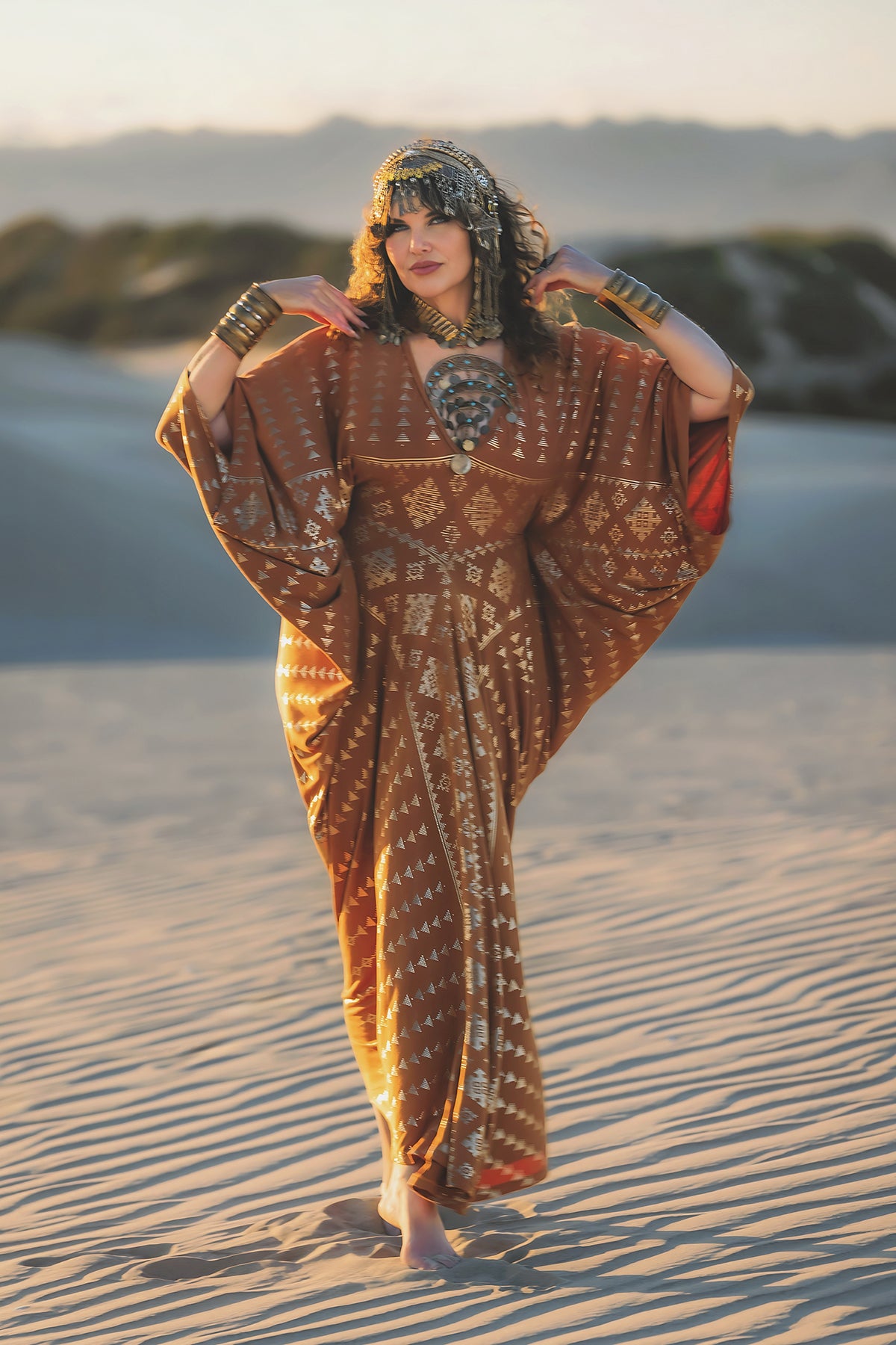 A woman stands barefoot on sand, sunlight illuminating her relaxed silhouette in the Melodia Designs Karnak Contessa Caftan 2.0 Copper, accented by gold accessories and a beaded headpiece, with blurred rocks in the background.
