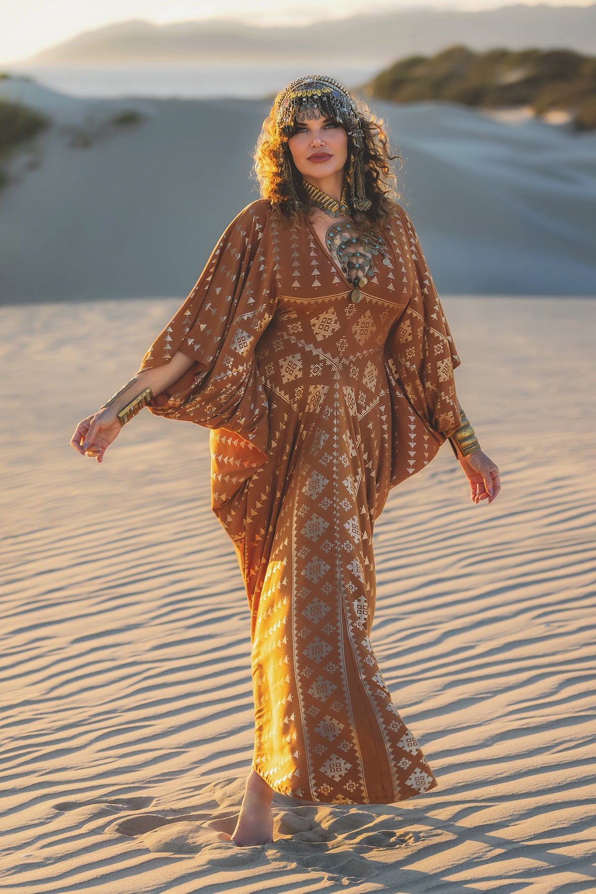 A woman in the Karnak Contessa Caftan 2.0 Copper by Melodia Designs strolls barefoot on sandy dunes at sunset, her batwing sleeves flowing and ornate headpiece shining against sunlit hills.