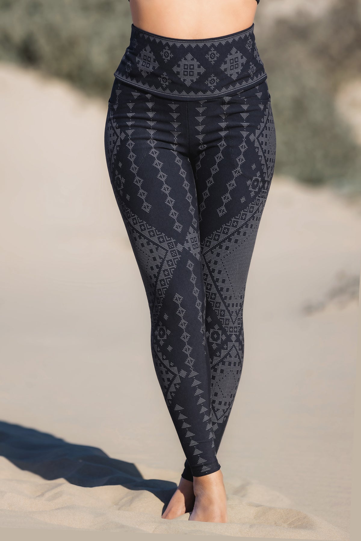 A person stands barefoot on sand, wearing Melodia Designs' Karnak Elsa Pocket Legging Shadow—high-waisted black leggings with a gray geometric pattern. The eco-luxe fabric is shown cropped chest-down against a blurred natural background.