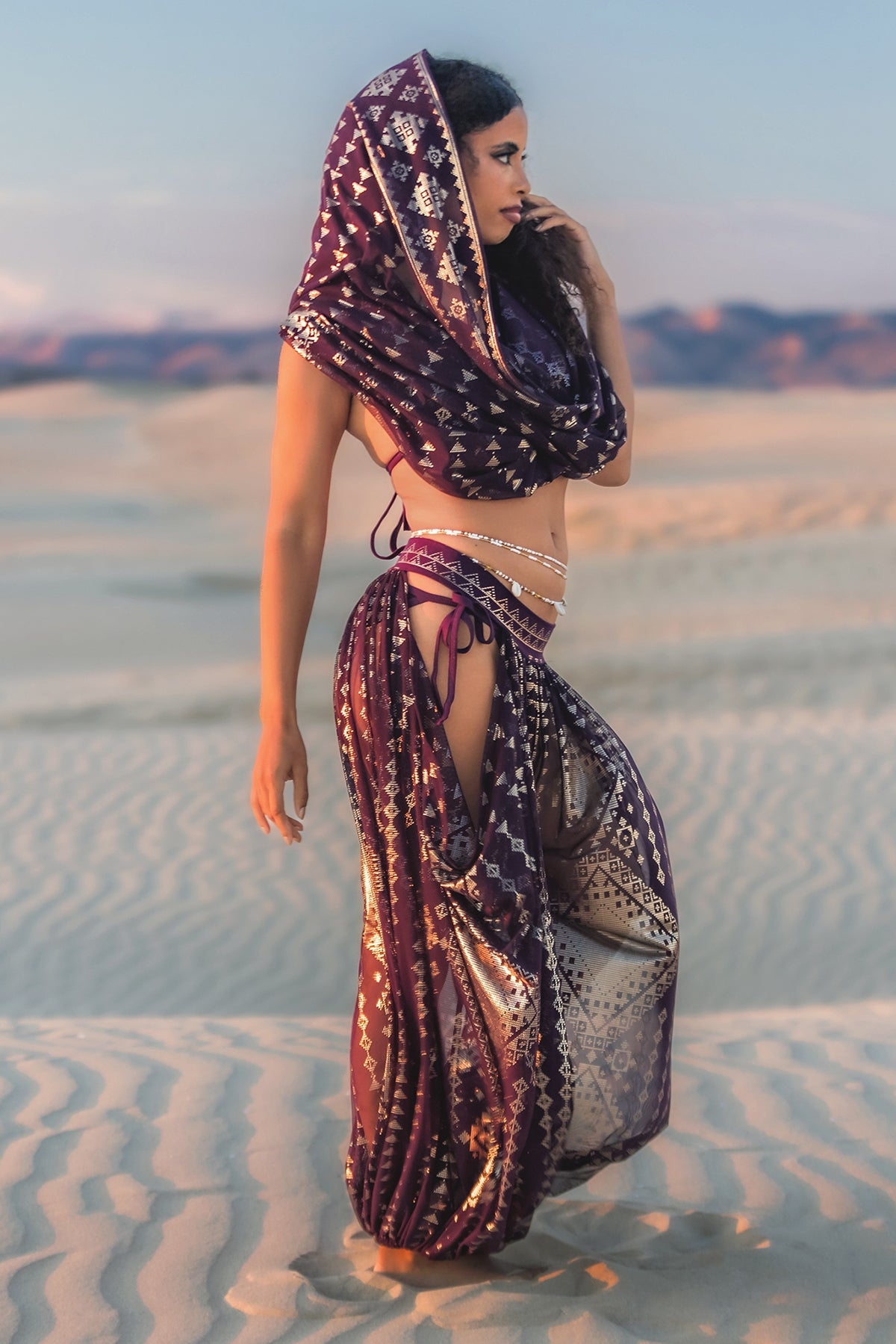 A woman poses on sandy dunes, wearing the Karnak Infinity Scarf in Aubergine-Gold by Melodia Designs—its hooded, flowing silhouette makes a striking statement against the serene desert backdrop.