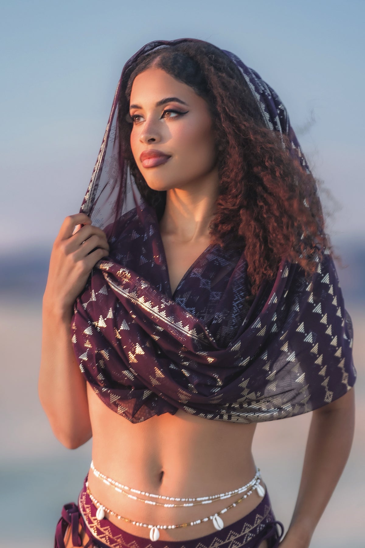 A woman with curly hair wears the Melodia Designs Karnak Infinity Scarf in Aubergine-Gold draped over her head and shoulders, styled with a shell belt and posing outdoors against a softly blurred background—a stunning statement piece.