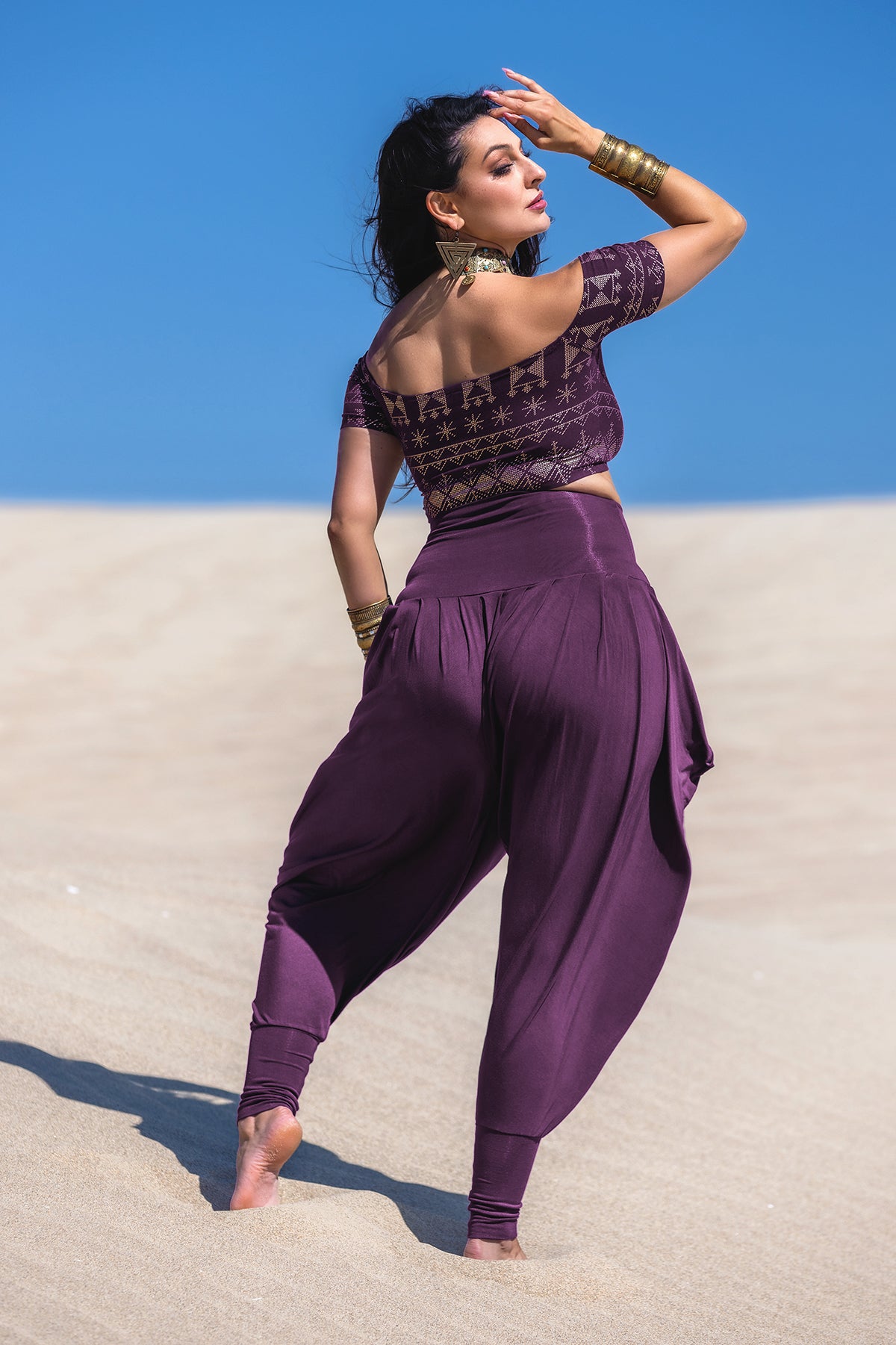 A woman poses barefoot on sandy dunes under a clear sky, wearing Melodia Designs’ Jazmine Harem Pant in Aubergine with a patterned crop top, gold bangles, and a choker—one hand on her head, the other on her hip.