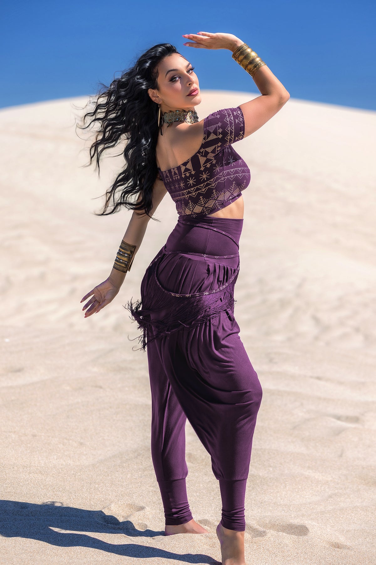 A woman models Melodia Designs’ Jazmine Harem Pant in Aubergine, posing gracefully on desert sand with gold bangles, a choker, and flowing wavy black hair, one arm raised and the other bent.