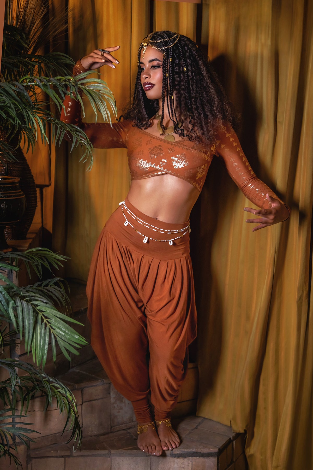 A woman with curly hair stands barefoot near plants and golden curtains, wearing the Jazmine Harem Pant Copper by Melodia Designs, paired with a sheer crop top and gold jewelry.