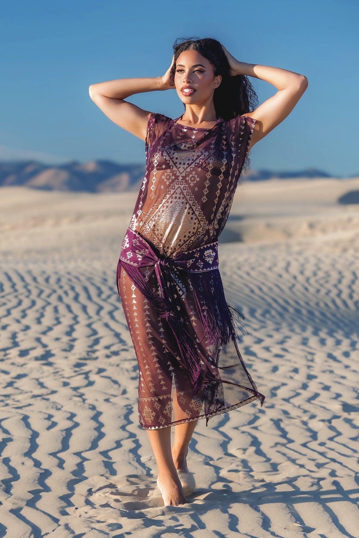 Woman in a sheer, patterned dress standing in a desert landscape with mountains in the background.