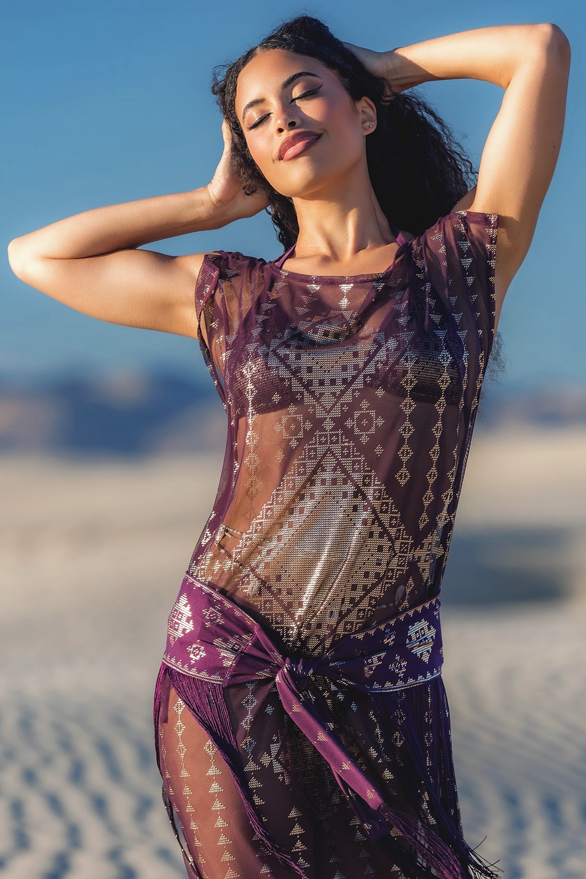 Woman in a patterned dress standing on a beach with a clear sky.