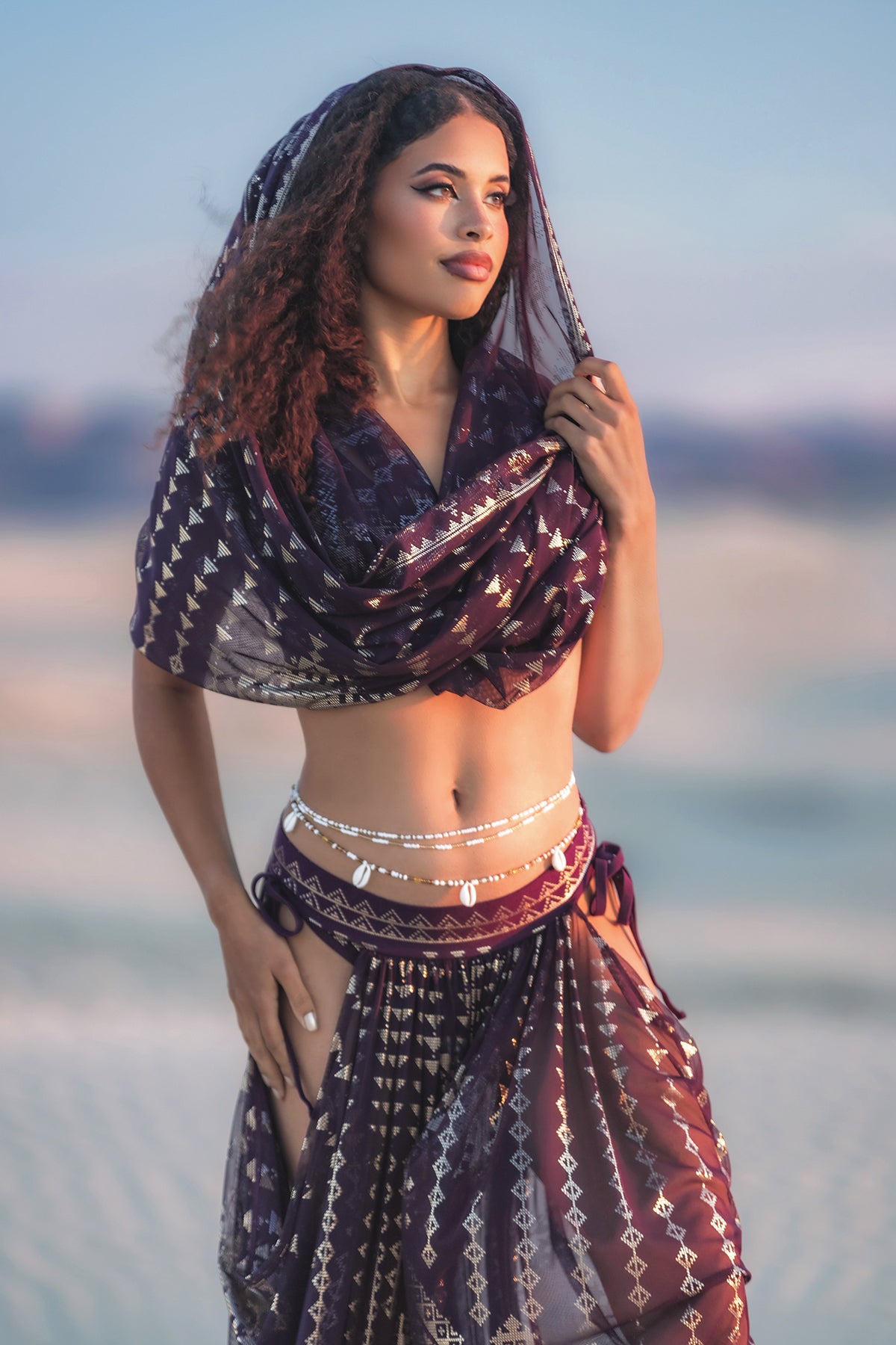 A woman stands on sand wearing Melodia Designs' Karnak Sahara Peekaboo Pantaloon in Aubergine-Gold with a matching headscarf and silver waist jewelry, gazing thoughtfully into the distance.