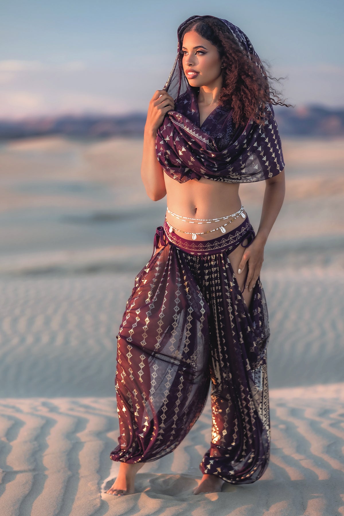 At sunset, a woman stands barefoot on sand dunes, wearing Melodia Designs’ Karnak Sahara Peekaboo Pantaloon in Aubergine-Gold and a scarf draped over her head, accessorized with silver jewelry at her waist and wrists.