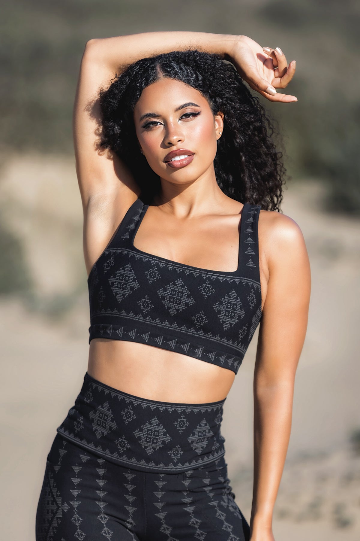 A woman with curly hair poses outdoors, wearing the Melodia Designs Karnak-Serena Bra 2.0 - Shadow and matching high-waisted leggings with a geometric pattern, one arm raised as she gazes confidently at the camera.