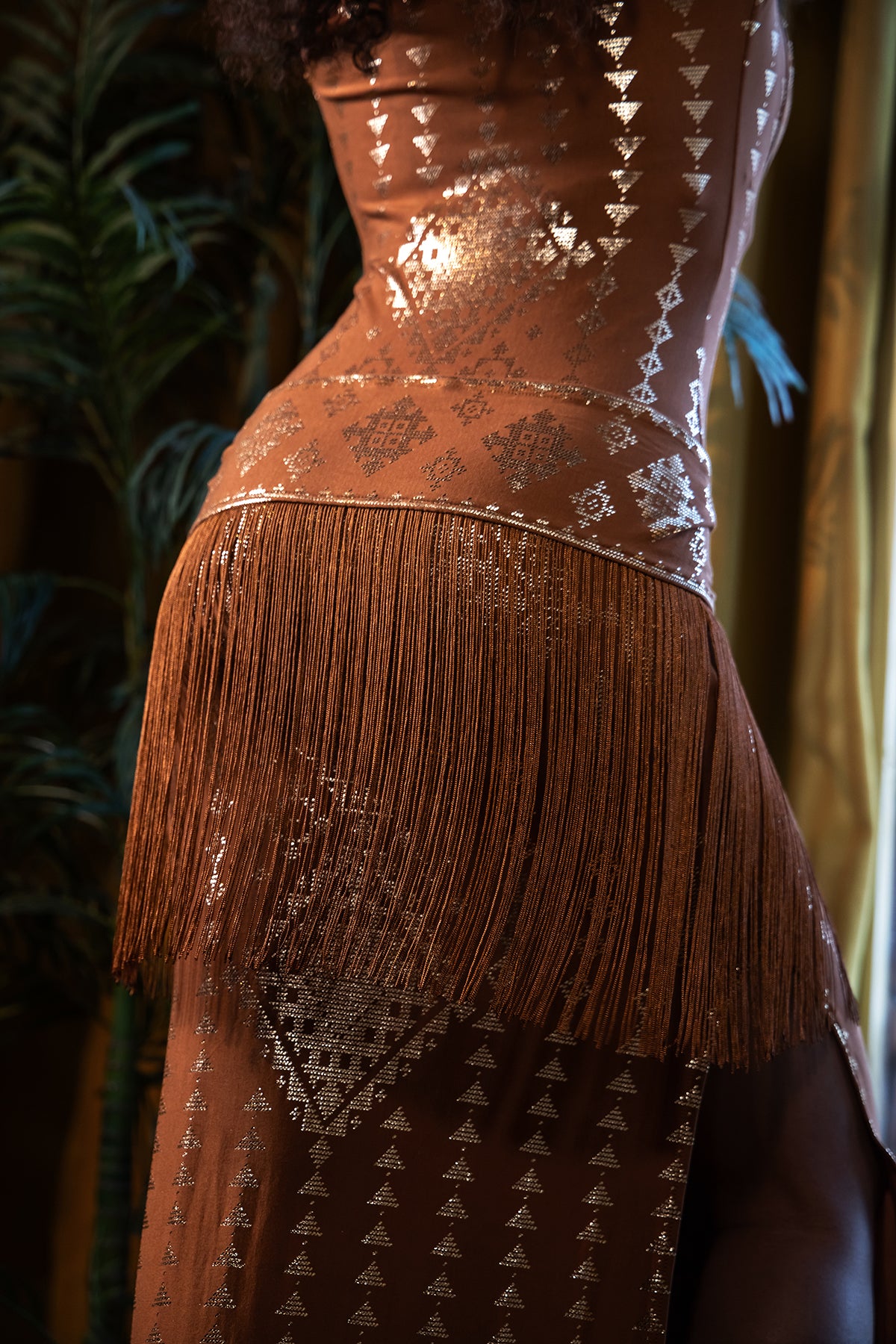A person wearing the Melodia Designs Karnak Shimmy Sash Copper-Gold, featuring metallic geometric patterns and chainette fringe at the hip, embraces resort style while standing beside a leafy green plant.