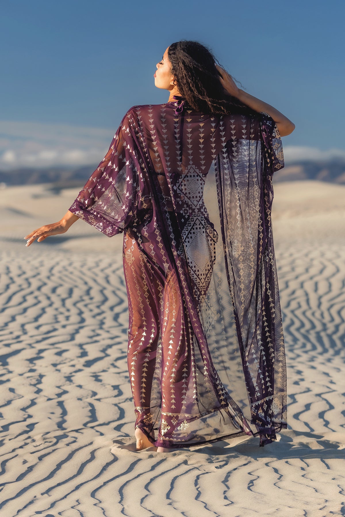 A woman stands barefoot on sandy dunes wearing the Melodia Designs Karnak Zsa Zsa Aubergine-Gold mesh duster with gold foil accents, arms outstretched and head tilted upward against clear skies and distant mountains.