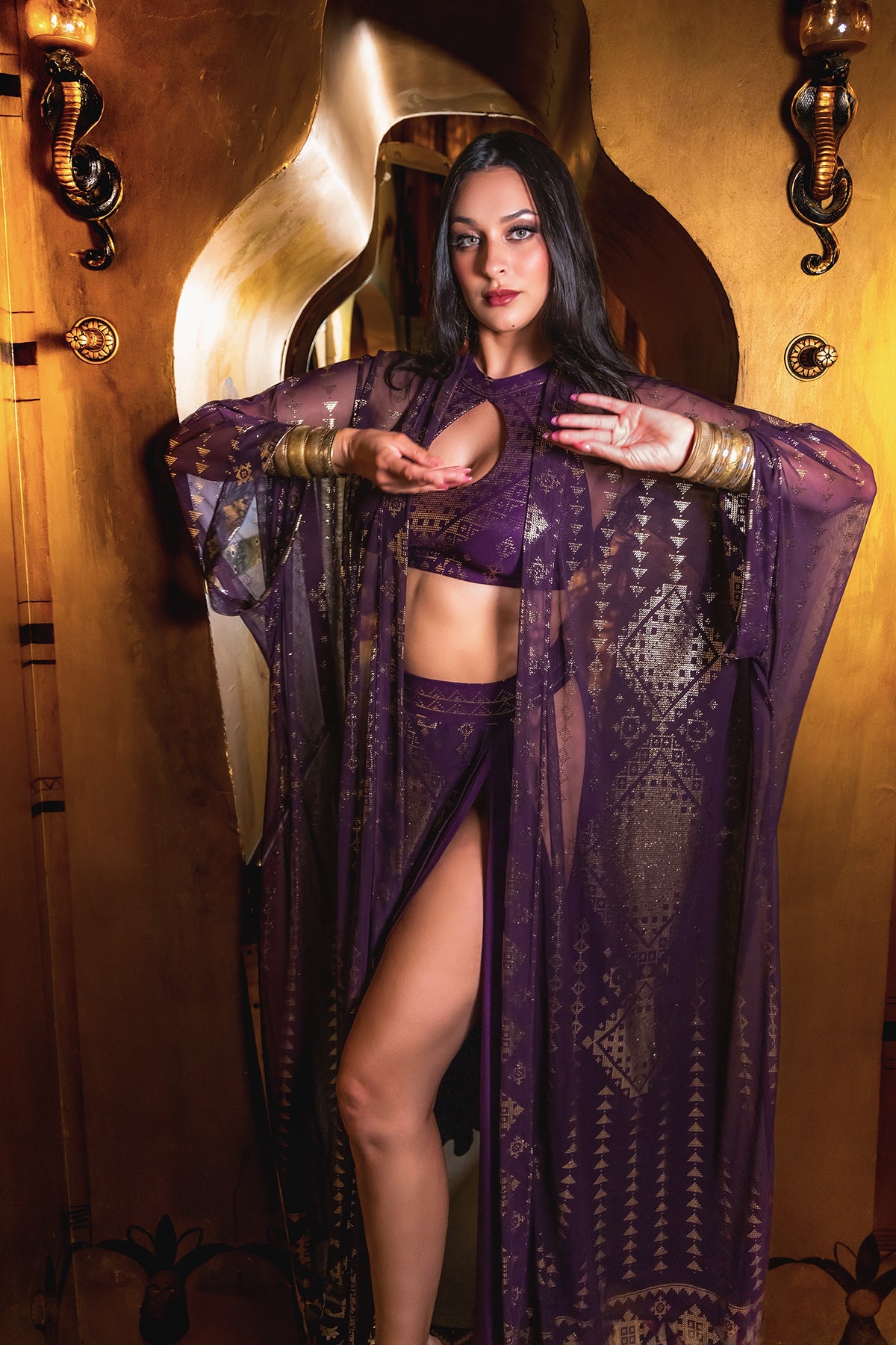 A woman poses in a gold setting, wearing the Melodia Designs Karnak Zsa Zsa Aubergine-Gold robe over matching lingerie, framed by a decorative arch and shimmering gold foil accents, with one leg forward and arms raised.
