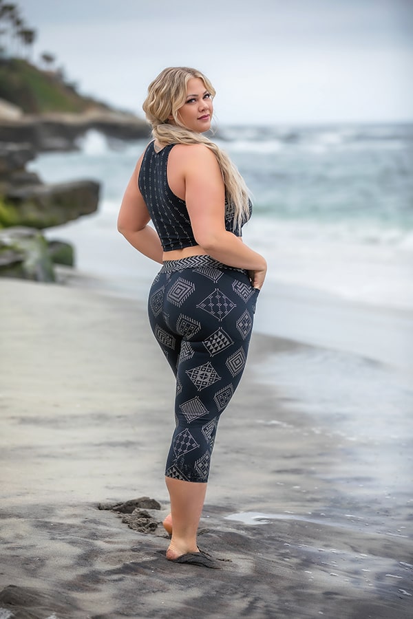 A woman in the Melodia Designs Laura Crop stands barefoot on a sandy beach, gazing over her shoulder at the camera. She wears patterned high-performance athletic wear, with ocean waves and rocky cliffs beneath an overcast sky.