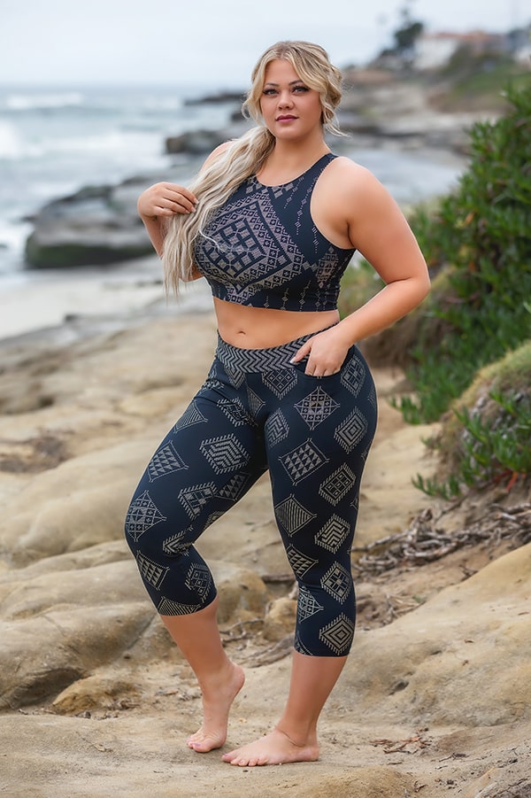 A woman with long blonde hair in a ponytail stands barefoot on rocky terrain by the ocean, wearing the Laura Crop by Melodia Designs—a navy blue and beige geometric-patterned athletic set made from high-performance fabric. She gazes ahead, one hand on her hip.